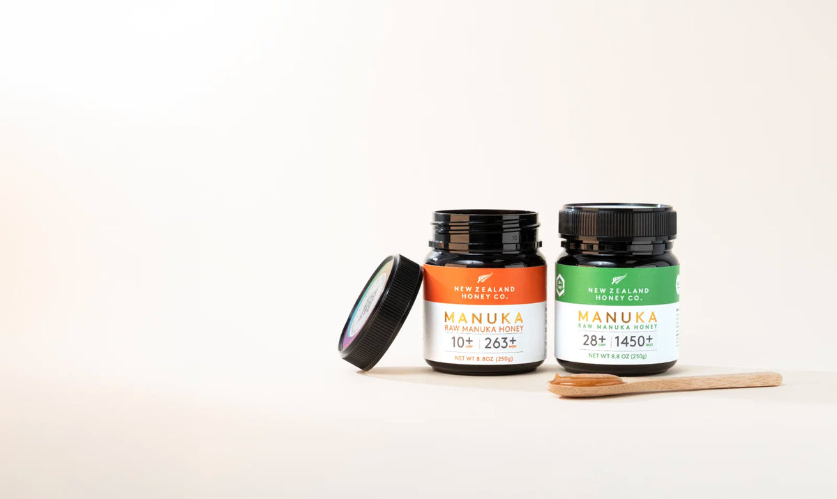 Two jars of New Zealand manuka honey beside a wooden spoon holding honey, with one lid leaning nearby.