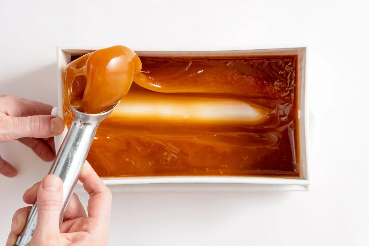 Hand holding a spoon with caramel above a rectangular container of melted caramel sauce.