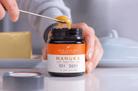 Hand lifting honey on a metal stick from an open UMF 10+ MGO 263+ Manuka honey jar
