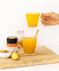 Hand pouring turmeric tea beside a UMF 10+ MGO 263+ Manuka honey jar with lemon and ginger