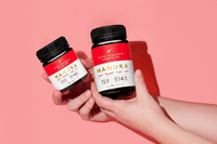Two UMF 15+ MGO 514+ Manuka honey jars held up against a pink background