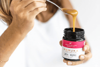 Hand lifting honey on a spoon from an open UMF 20+ MGO 829+ Manuka honey jar