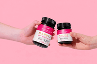 Two hands holding UMF 20+ MGO 829+ Manuka honey jars against a pink backdrop