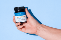 Hand holding a UMF 26+ MGO 1282+ Manuka honey jar against a blue background