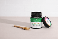 Open UMF 28+ MGO 1450+ Manuka honey jar with bamboo spoon and lid on a pink surface